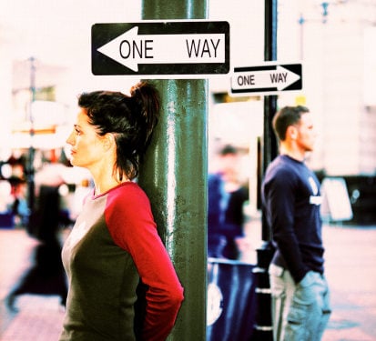 side profile of a woman in the foreground and a man in the background both leaning against green posts with one way traffic signs above their head with each sign facing the opposite direction