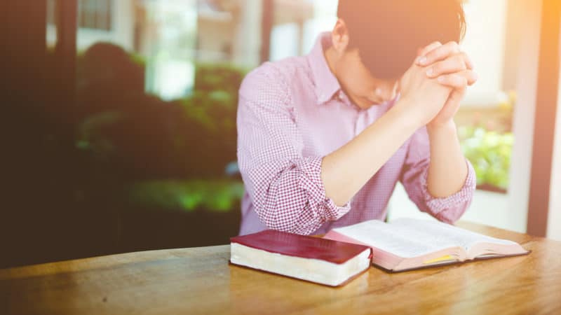 young man sitting at table with open scriptures saying a prayer before beginning bible study