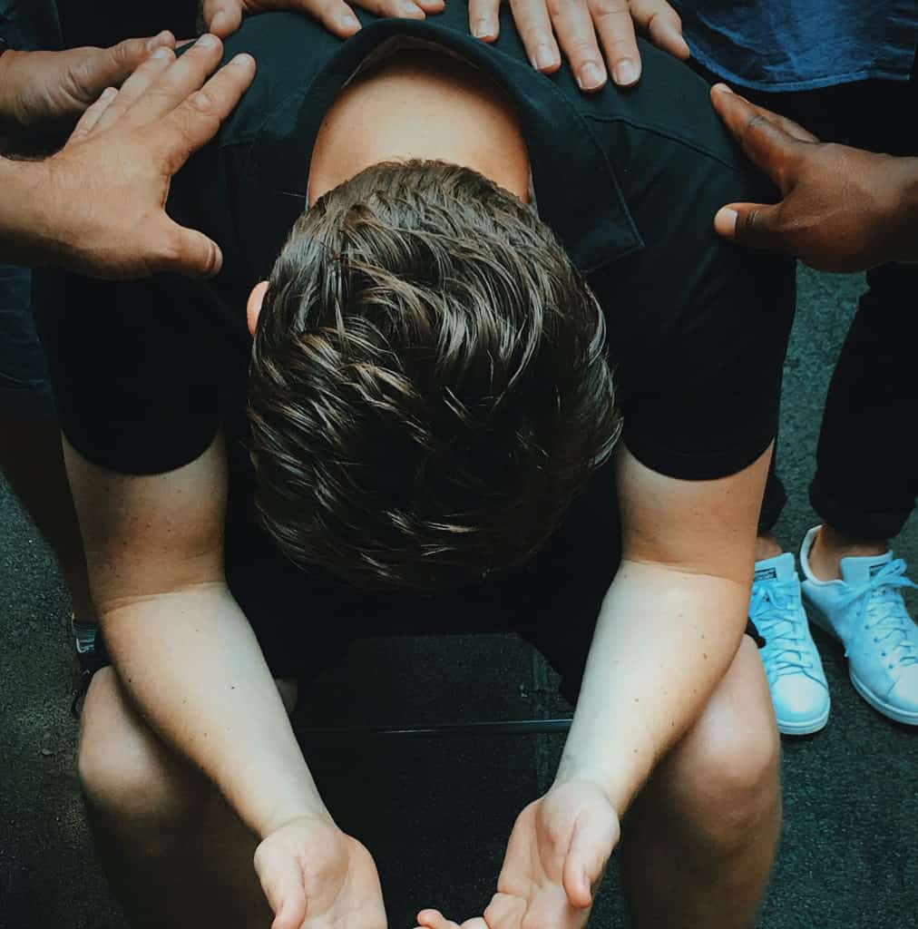 young man sitting bent over on chair with hands of friends resting on him in prayer
