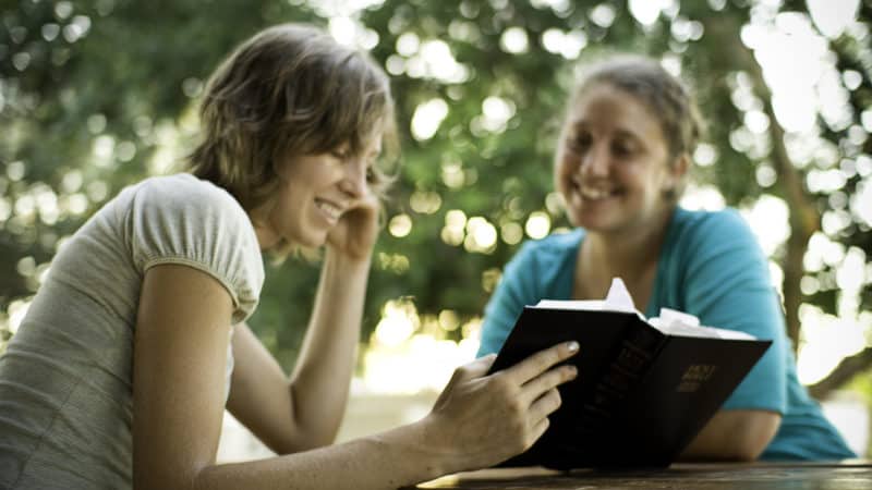 two women sitting outdoors at a picnic table reading the bible together to better understand biblical principles of living