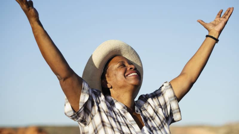 happy woman older woman wearing a straw hat and grey and white plaid shirt out in a field with arms raised feeling happy about the gift of salvation through Jesus