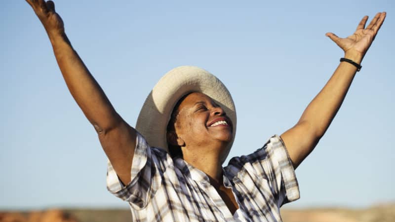 older woman wearing a straw hat and grey and white plaid shirt out in a field with arms raised feeling happy about the gift of salvation through Jesus