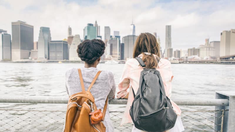 two women with backpacks standing on pier looking at cityscape