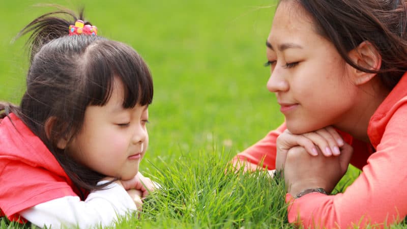 mother and young daughter lying on their stomachs on the grass facing each other with their eyes closed