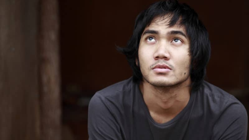 young man with dark hair sitting in front of an old building looking up into the sky contemplating salvation