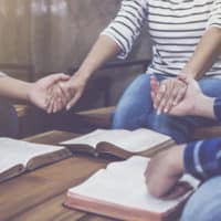 Three friends sitting in front of thier own bibles with their hands joined in prayer