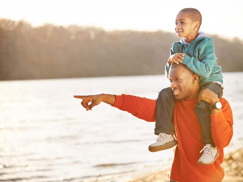 Father carrying his little boy on his shoulders on a beach by a lake, pointing at the difference things God created.