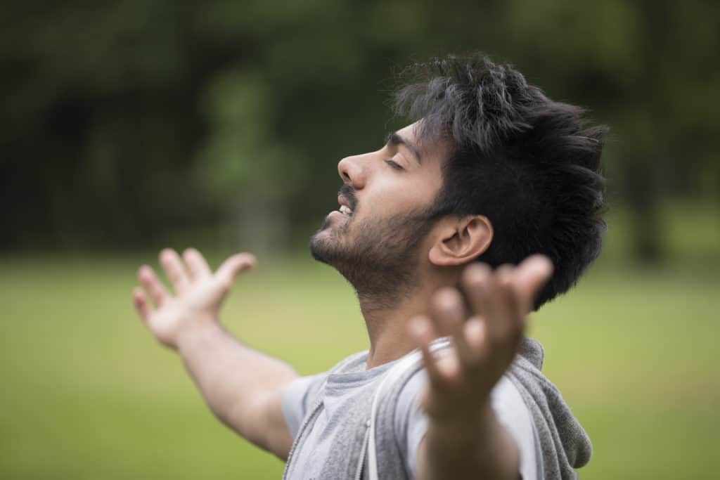 Asian man freely worshiping God on Saturday outdoors with his arms raised to the heavens.