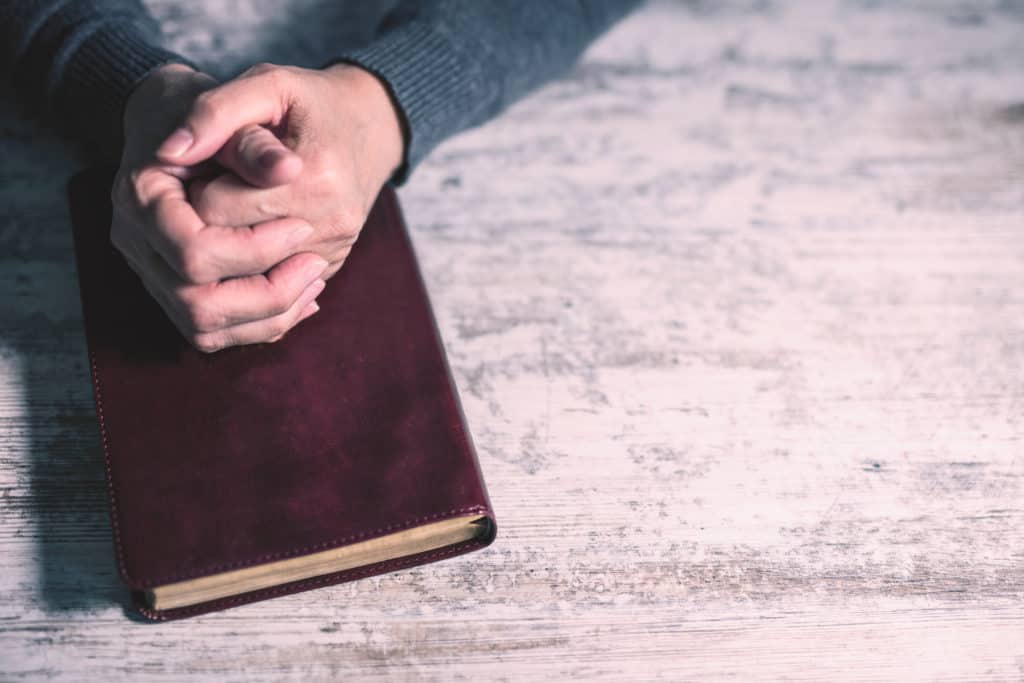 Hands folded on a closed brown Bible, praying before studying Scripture.