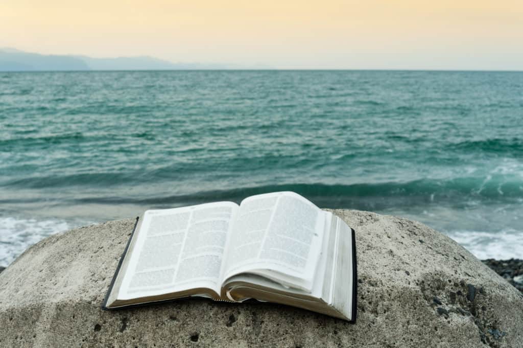 Open bible on top of a rock in front of the sea of greenish waters and an orange sky. 
