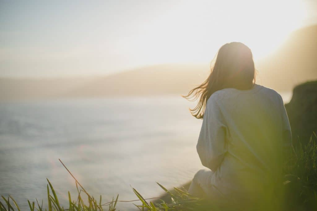 A woman sitting on the grass overlooking an ocean, contemplating creation.