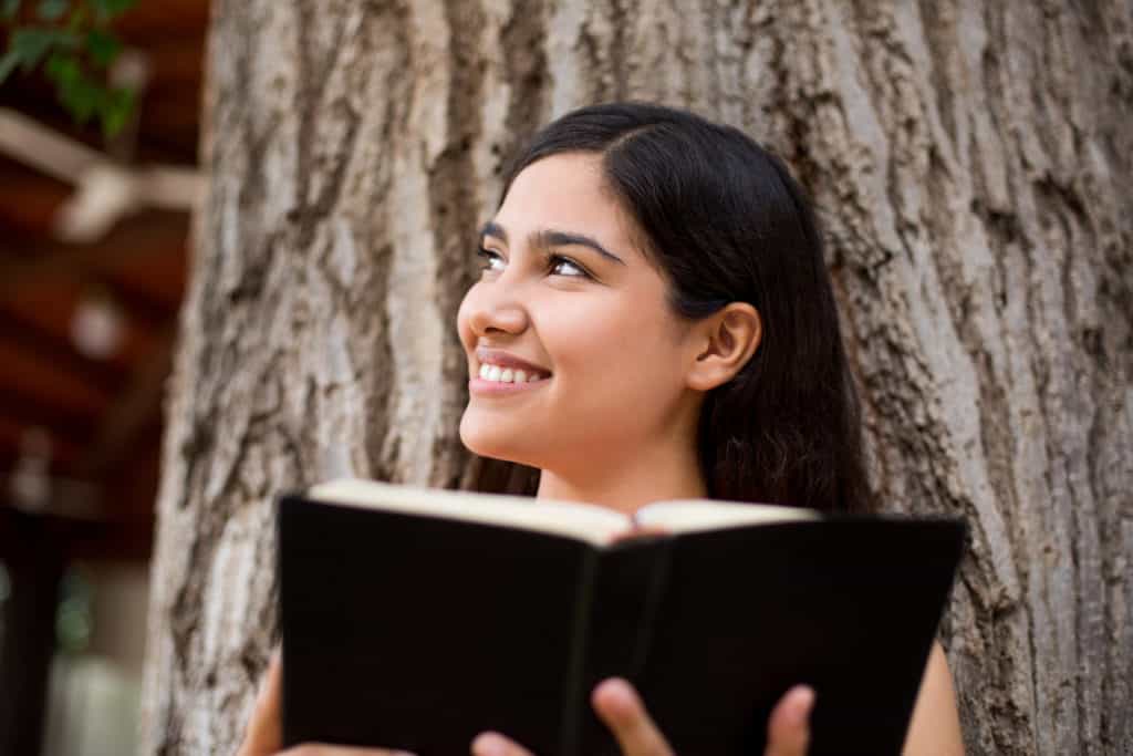 Young Hispanic woman smiling and enjoying Sabbath while sitting against a tree holding an open black Bible.