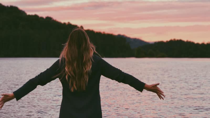 Woman stands in front of lake at sunset with her arms outstretched enjoying nature.