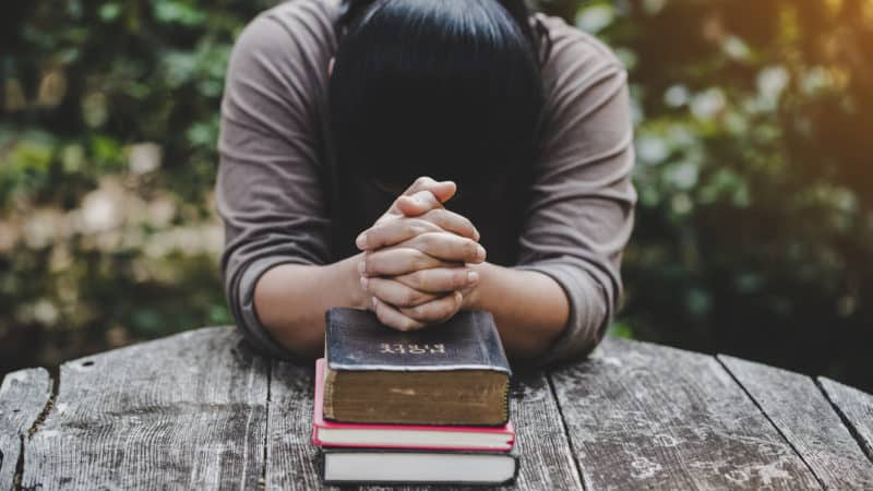 Person with head bowed and hands folded in prayer on a Holy Bible