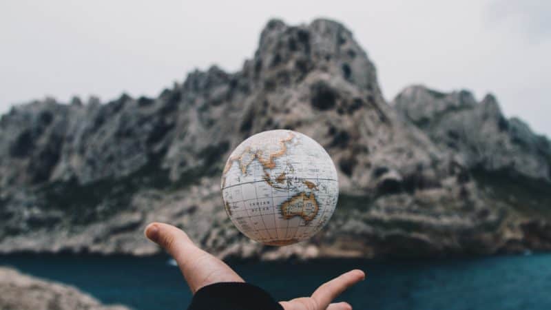 a person tossing a small globe above their hand with ocean rock formations and an inlet blurred in the distance