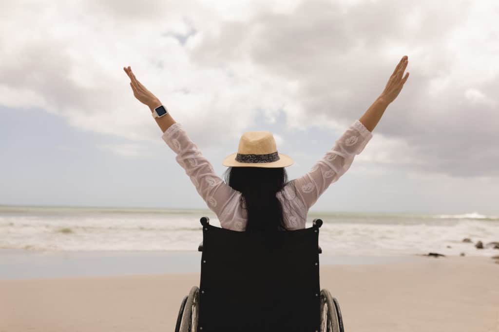 Rear view of a woman in a wheel chair with arms stretched out at beach in the sunshine