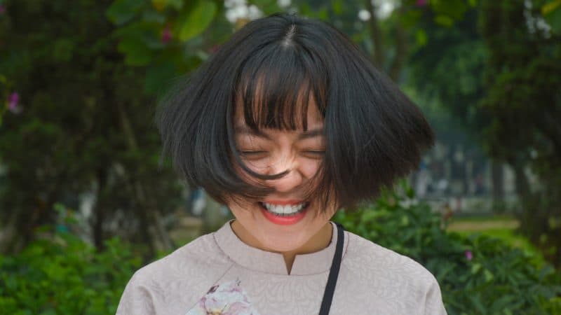 a happy woman with a big smile standing outdoors with shoulder length black hair blowing in the breeze