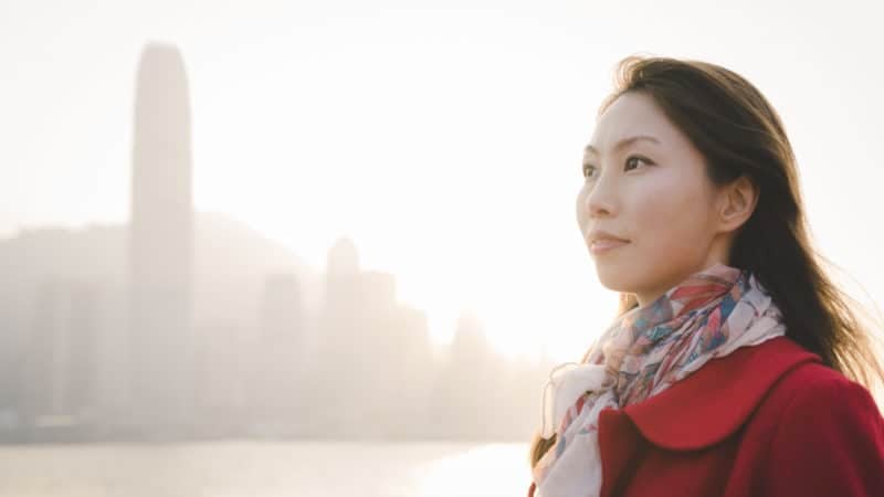 Woman in a red wool coat and scarf standing with a skyline in the background thinking about the gift of salvation