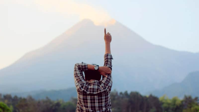 A man standing in front of a mountain with this hand raised and finger pointed towards the sky.