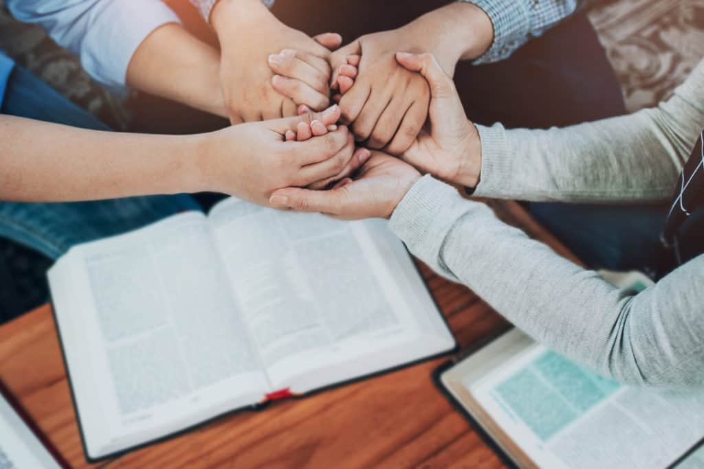 Christian friends joining hand and pray together over Bible on wooden table