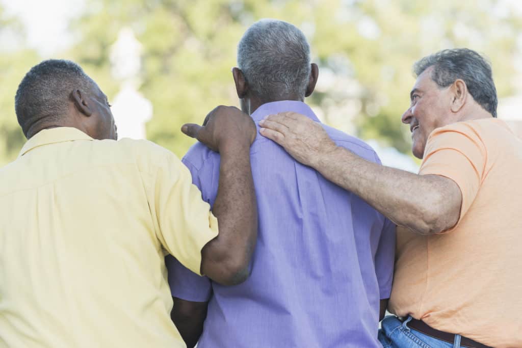 Rear view of three senior men sitting together on a park bench. The two men on the ends seem to be comforting their friend sitting between them.