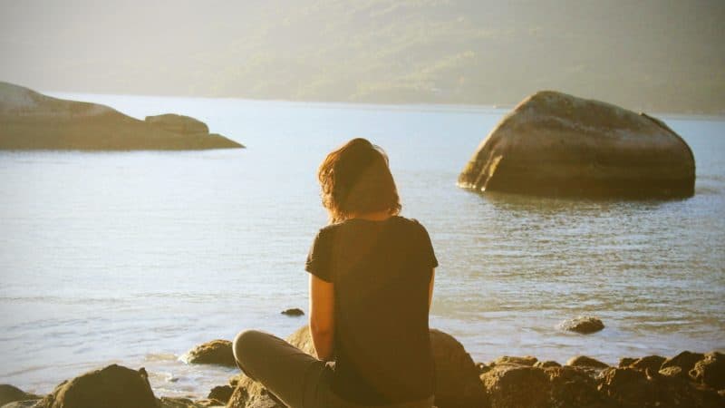 A woman sitting on a rock by a body of water at sunset thinking about the goodness of God and His love