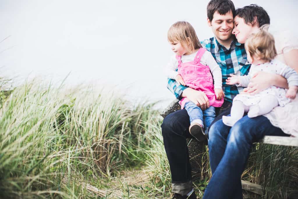 A young peaceful family relax and smile together as they sit with their two children on a park bench. 