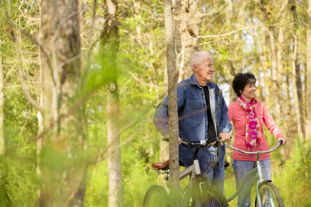 Active senior couple outside riding bikes in a wooded park area of their neighborhood.