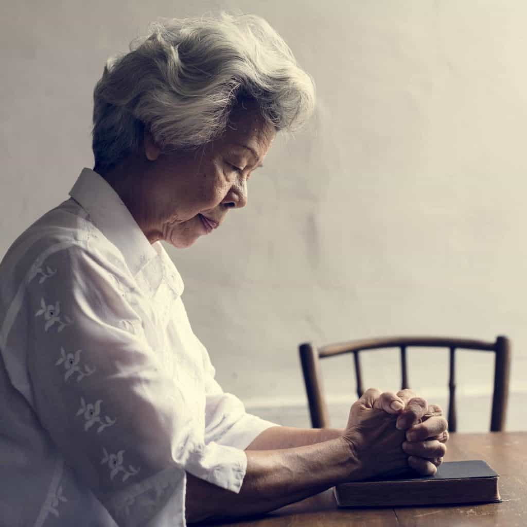 A woman praying with her Bible at a table as we pray that God will help us to understand the prophecies in the Bible