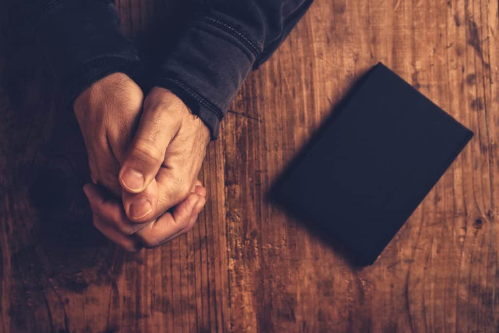 overhead view of a man's praying hands and a black bible beside him on a wooden desk as he contemplates the gift of prophecy