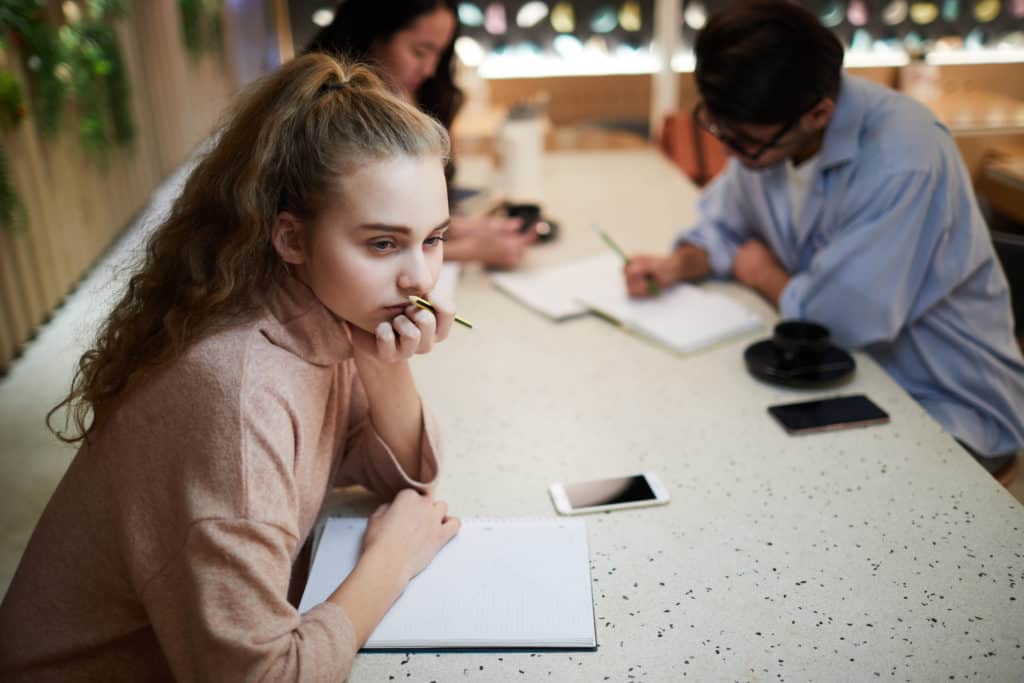 Pensive teenage girl sitting at table over open notebook 