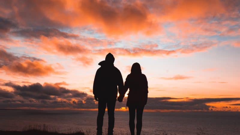 Silhouette of two people holding hands looking out over water at sunset.