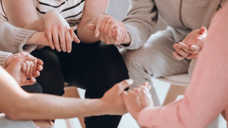 A group of people sitting in a circle holding hands in prayer during a prayer meeting session as we meet in small groups