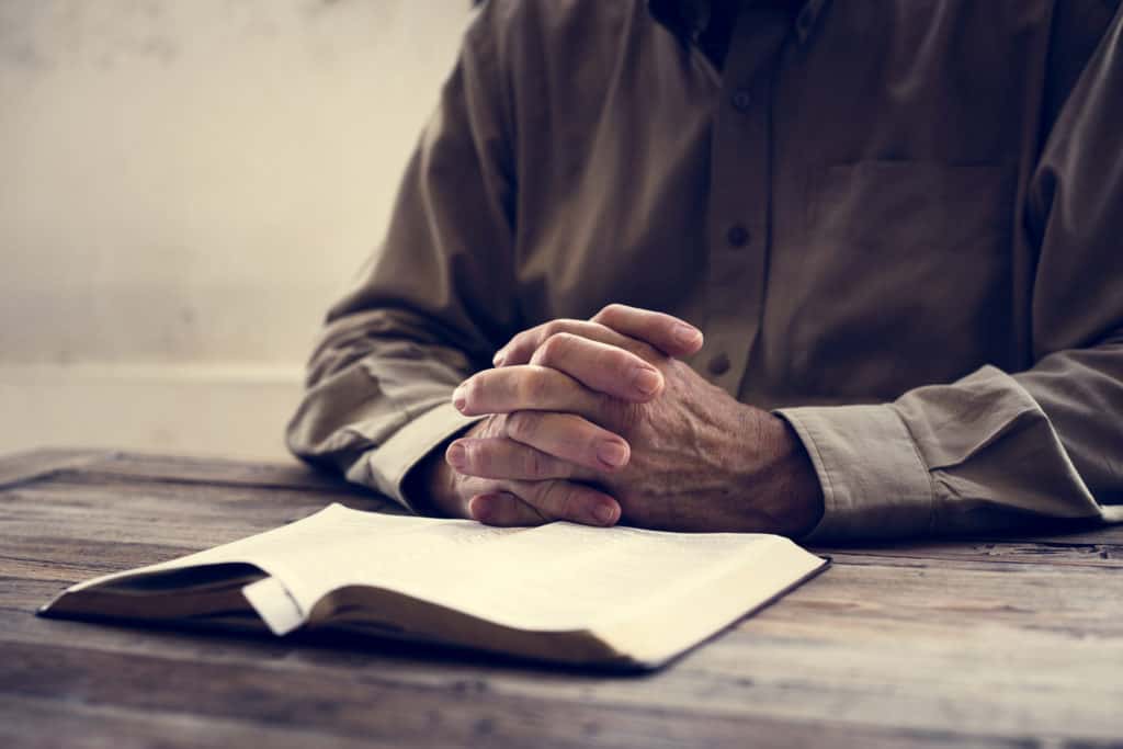 An old man reading the Bible with his hands folded in front of him. 