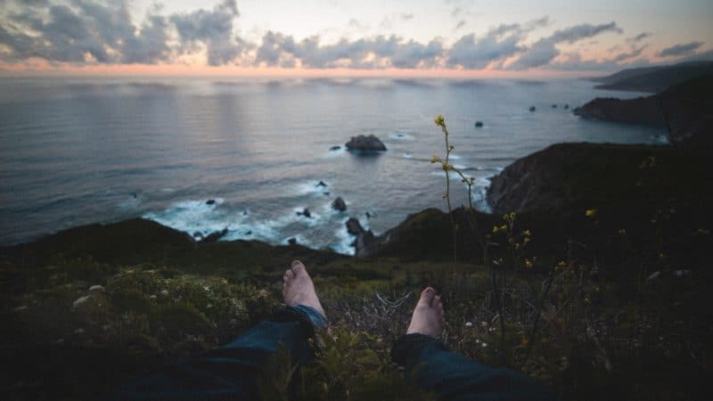 A person sitting on a grassy cliff looking out over the vast expanse of sea below and clouds above