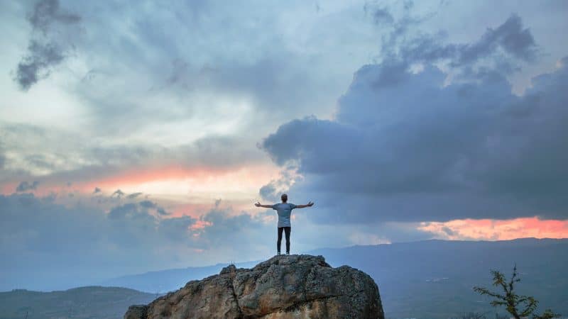A person standing on the top of a boulder looking at the cloudy sky with their arms outstretched, waiting for Jesus' return