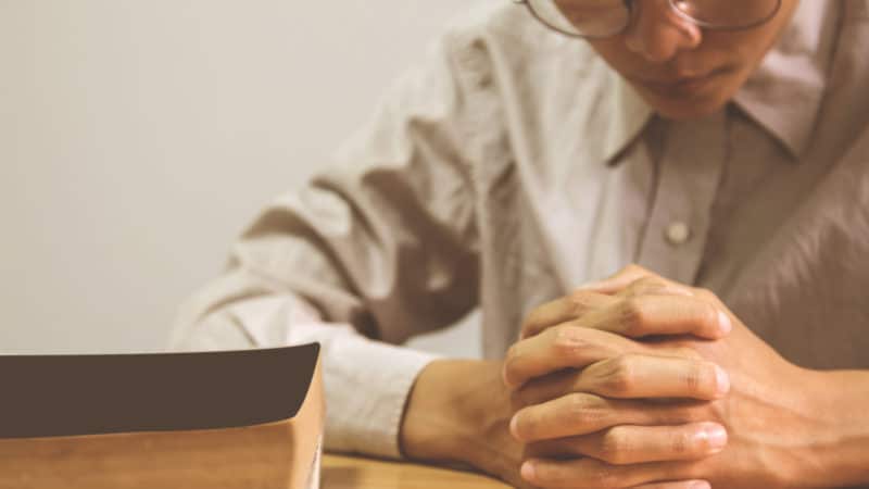 close up of a man praying with a Bible next to him on the table. 