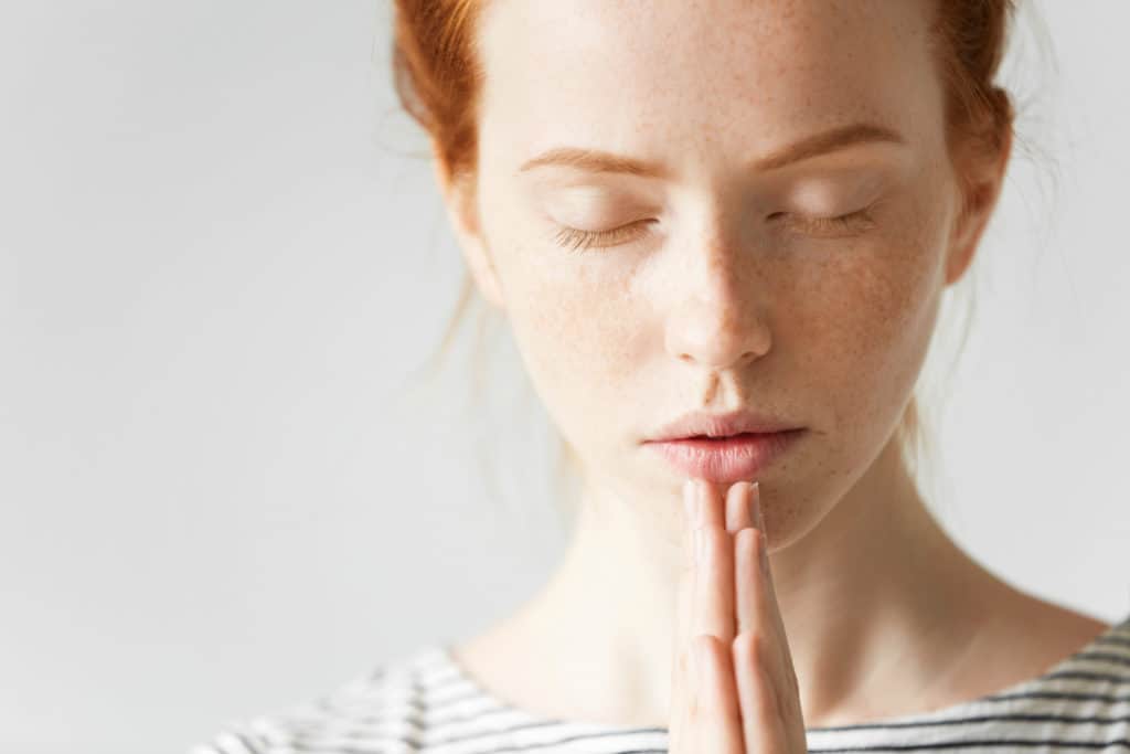 closeup of a young woman with read hair praying with her eyes closed and hands folded