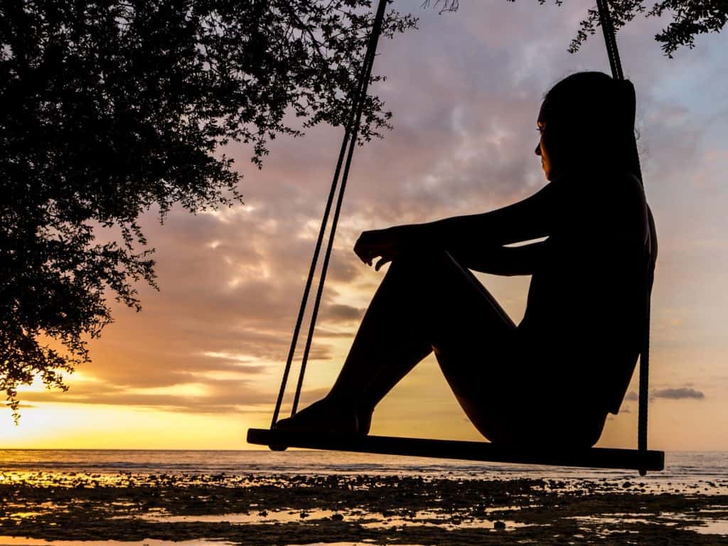 A woman sitting on a swing looking out over the water at sunset.