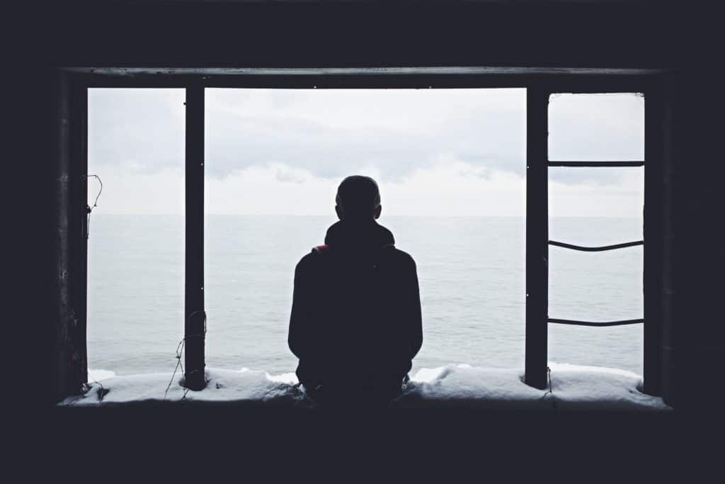 A person sitting on a window ledge in an abandoned building looking out at the ocean.