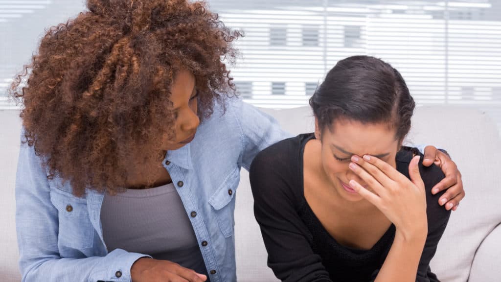 Sad woman crying next to her friend who is comforting and interceding for her.