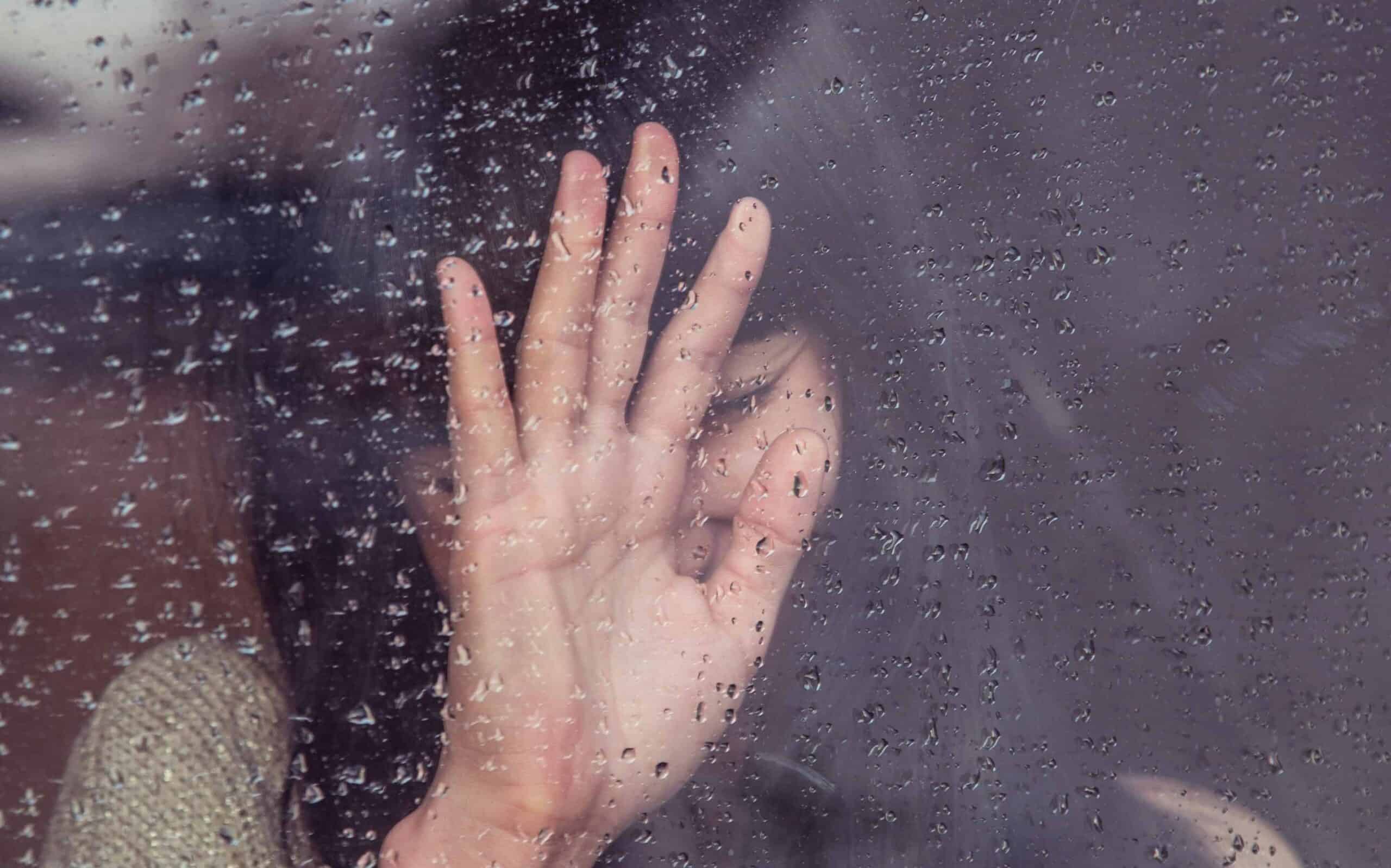 A sad, crying woman standing at a window with raindrops on it. 