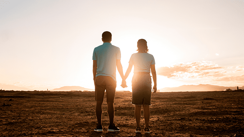 Two people holding hands while watching the sun rise in a desert like setting. 