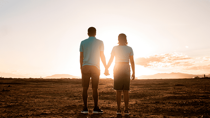 Two people holding hands while watching the sun rise in a desert like setting.