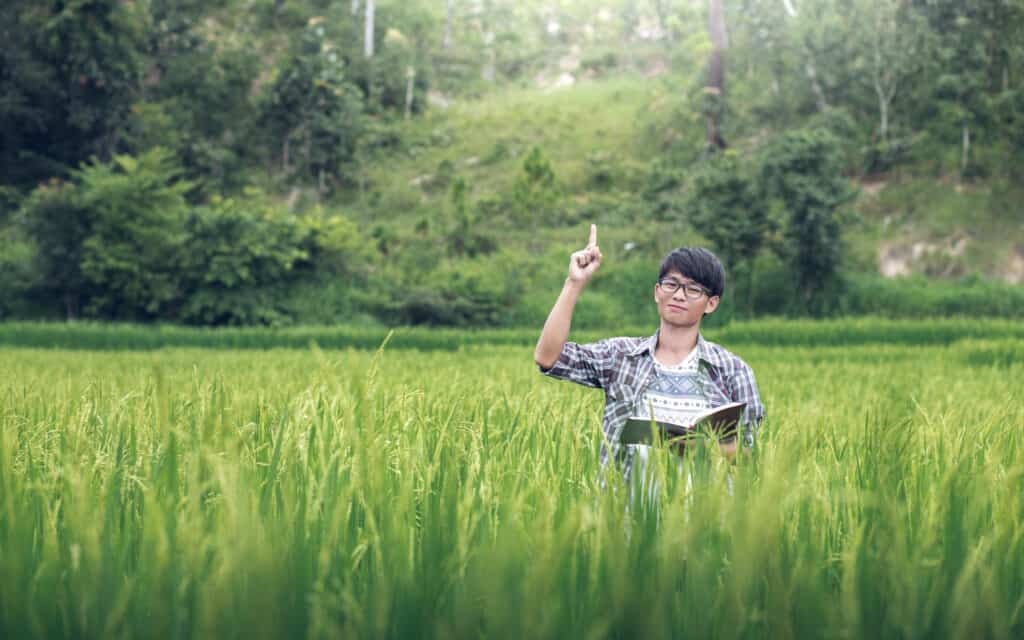 Man sitting in a field of grass looking at the camera smiling, holding the bible while pointing towards the sky. 