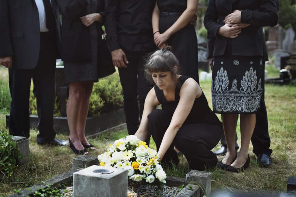 Woman laying flowers on the grave in front of her. A group of people dressed in black stand behind.