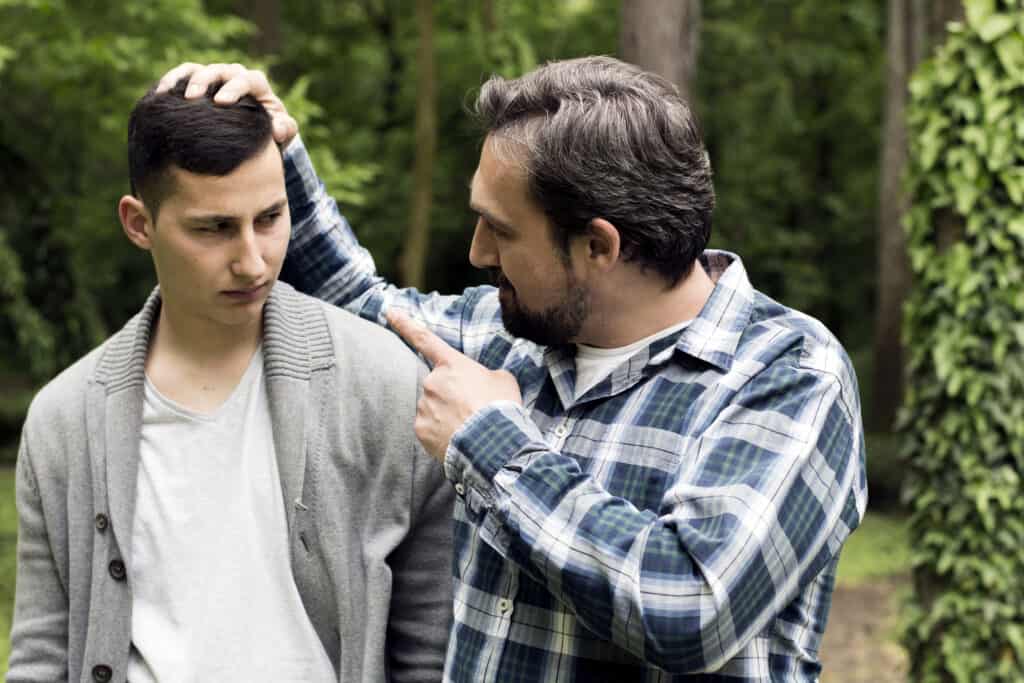 Two men stand close together one man is holding the others head and pointing in his face.