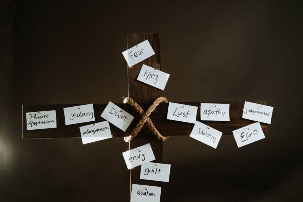 Wooden cross with words written on paper and the paper nailed to the cross. The words say, fear, lying, lust, apathy, judgmental, idolatry, ego, envy, guilt, isolation, doubt, unforgiveness, jealousy, passive aggression. 