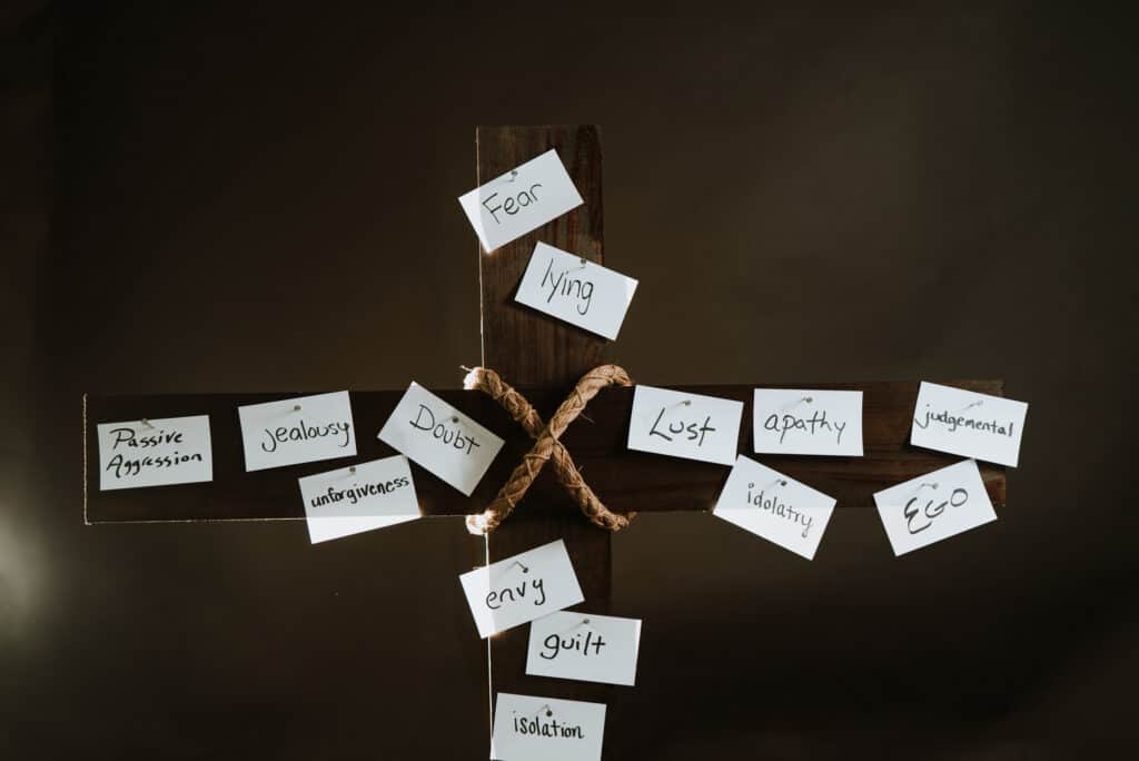Wooden cross with words written on paper and the paper nailed to the cross. The words say, fear, lying, lust, apathy, judgmental, idolatry, ego, envy, guilt, isolation, doubt, unforgiveness, jealousy, passive aggression.