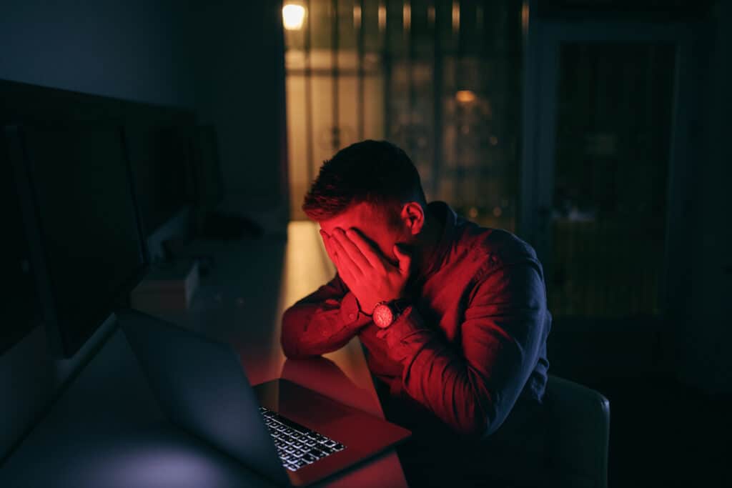 Man sitting in a dark room in front of a computer with a red light shining on him. His hands cover his face. 